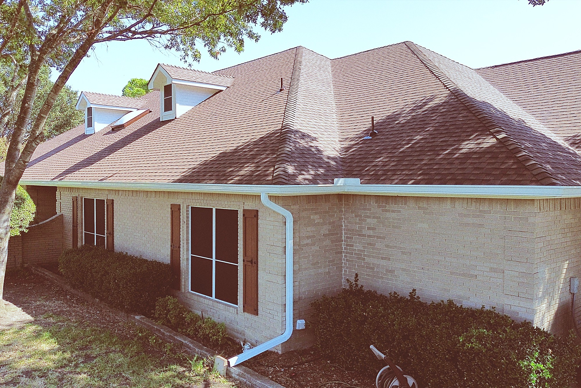 A brown shingle roof above a white gutter and downspout on a tan brick home