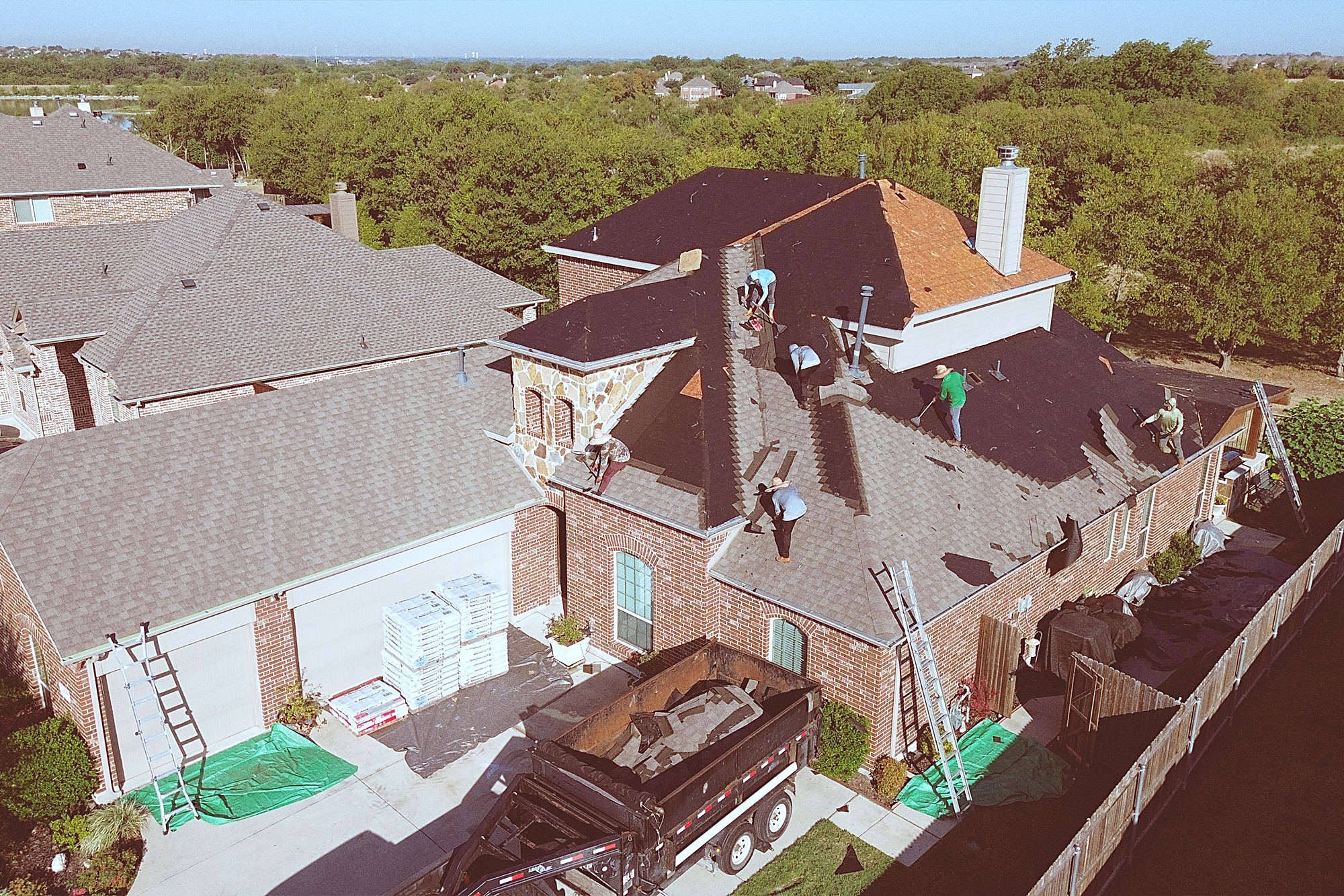 A crew shingling a two-story brick and stone home with a dump trailer in the drive